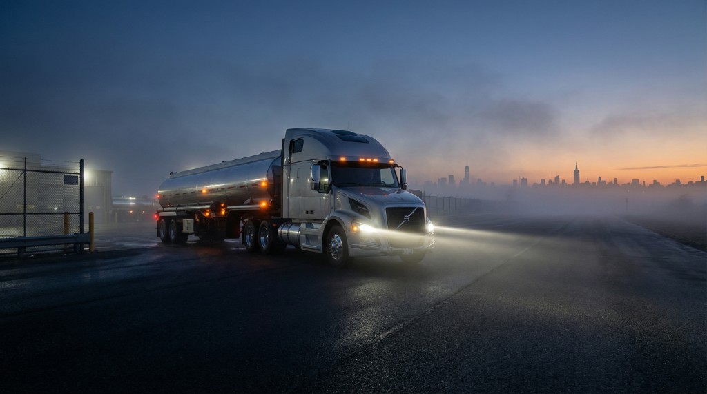 Silver tanker with headlights on in fog at dusk, city skyline in the distance